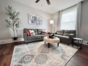 A living room with a grey sectional sofa, a round ottoman, a floor lamp, and a large window with curtains.
