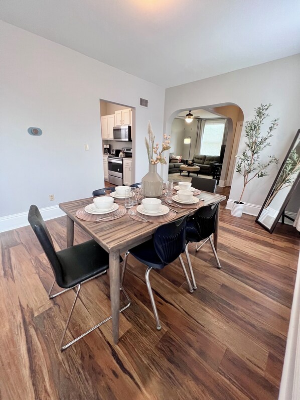 A dining area with a wooden table set for four, surrounded by black chairs. A kitchen is visible in the background.