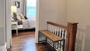 A room with a wooden bench, a bed with a striped bedspread, a nightstand with a lamp, and a window with curtains. - Spacious 3 Bedroom Sleeps 5 - Walk to Botanical Garden - Long Term Pet Friendly (St. Louis)