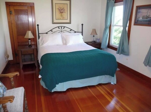 Un dormitorio con piso de madera, una cama con colcha verde, dos mesitas de noche con lámparas, una puerta de madera y una ventana con cortinas.