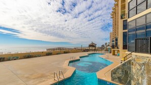Zona de piscina con vista a la playa, un quiosco y un edificio con grandes ventanales.