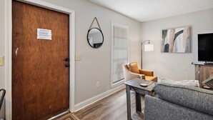 A living room with a wooden door, a mirror, a window with blinds, a sofa, a table, and a TV. - Cottage 2 - Bourbon Way (Ridgeland)