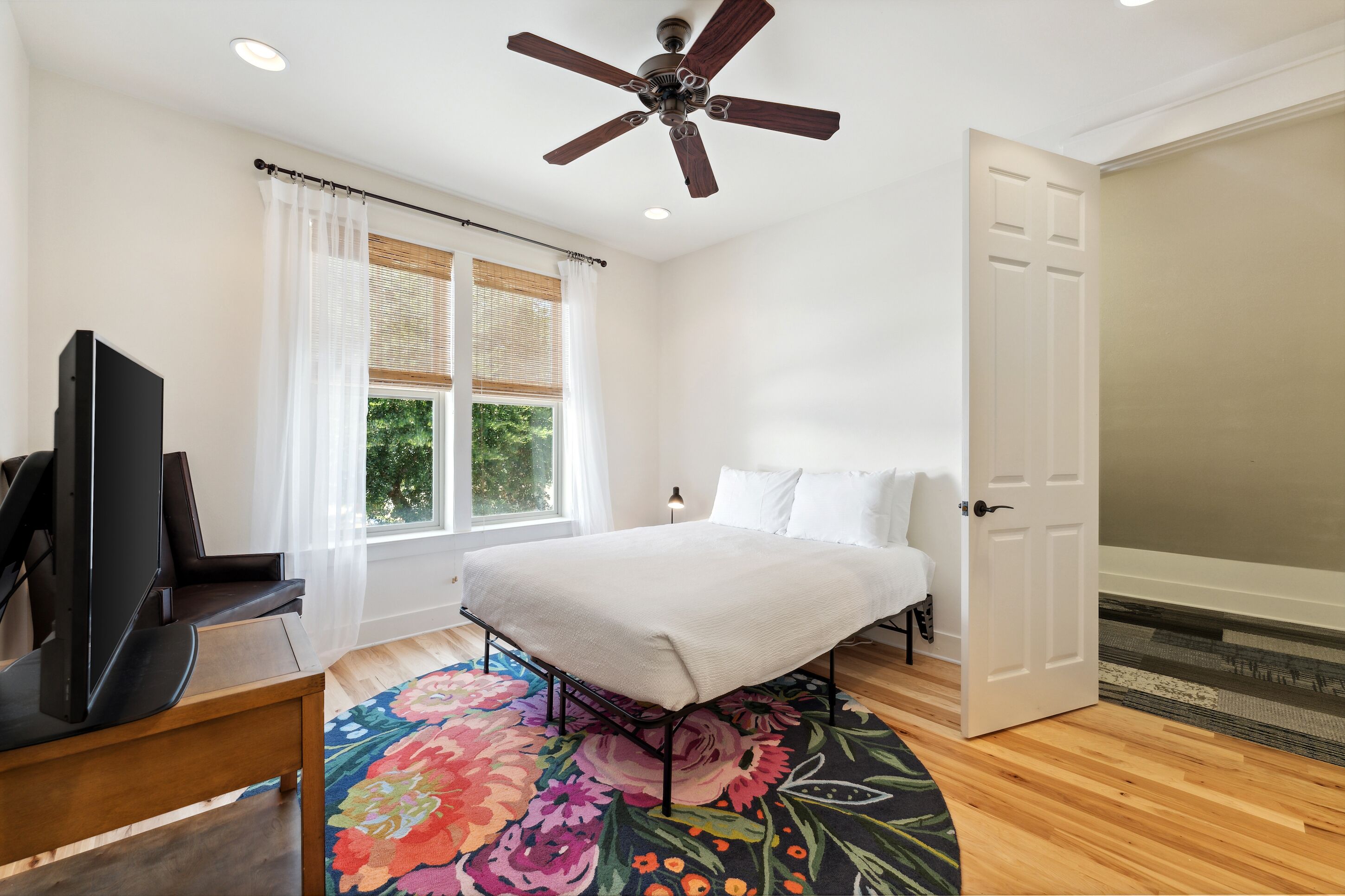 A bedroom with a bed, a television, a ceiling fan, a window with curtains, and a door.