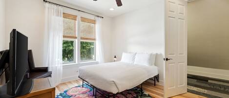 A bedroom with a bed, a television, a ceiling fan, a window with curtains, and a door.