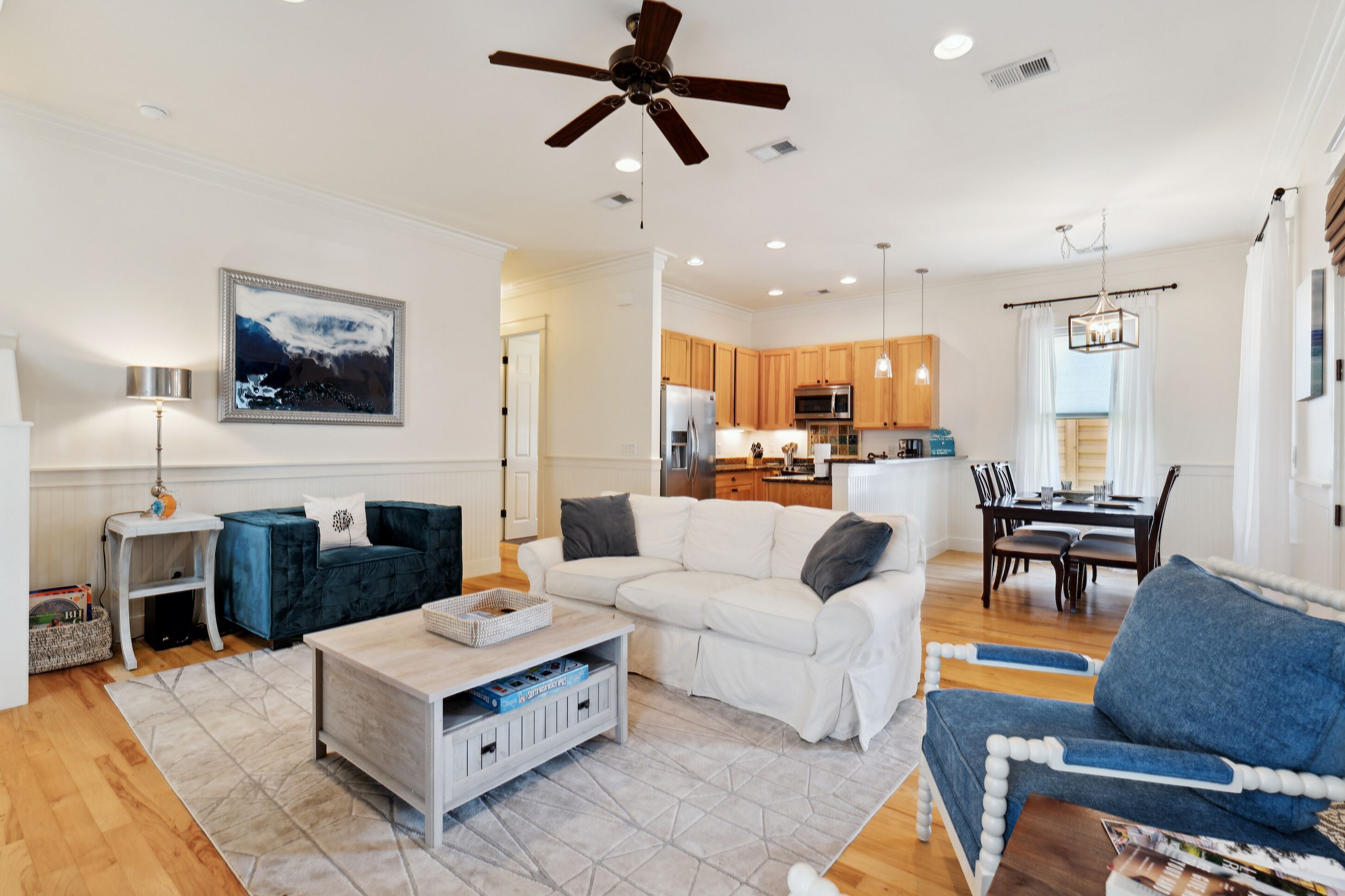 A spacious living room with a white sectional sofa, a blue armchair, and a coffee table. A ceiling fan and recessed lighting are visible.