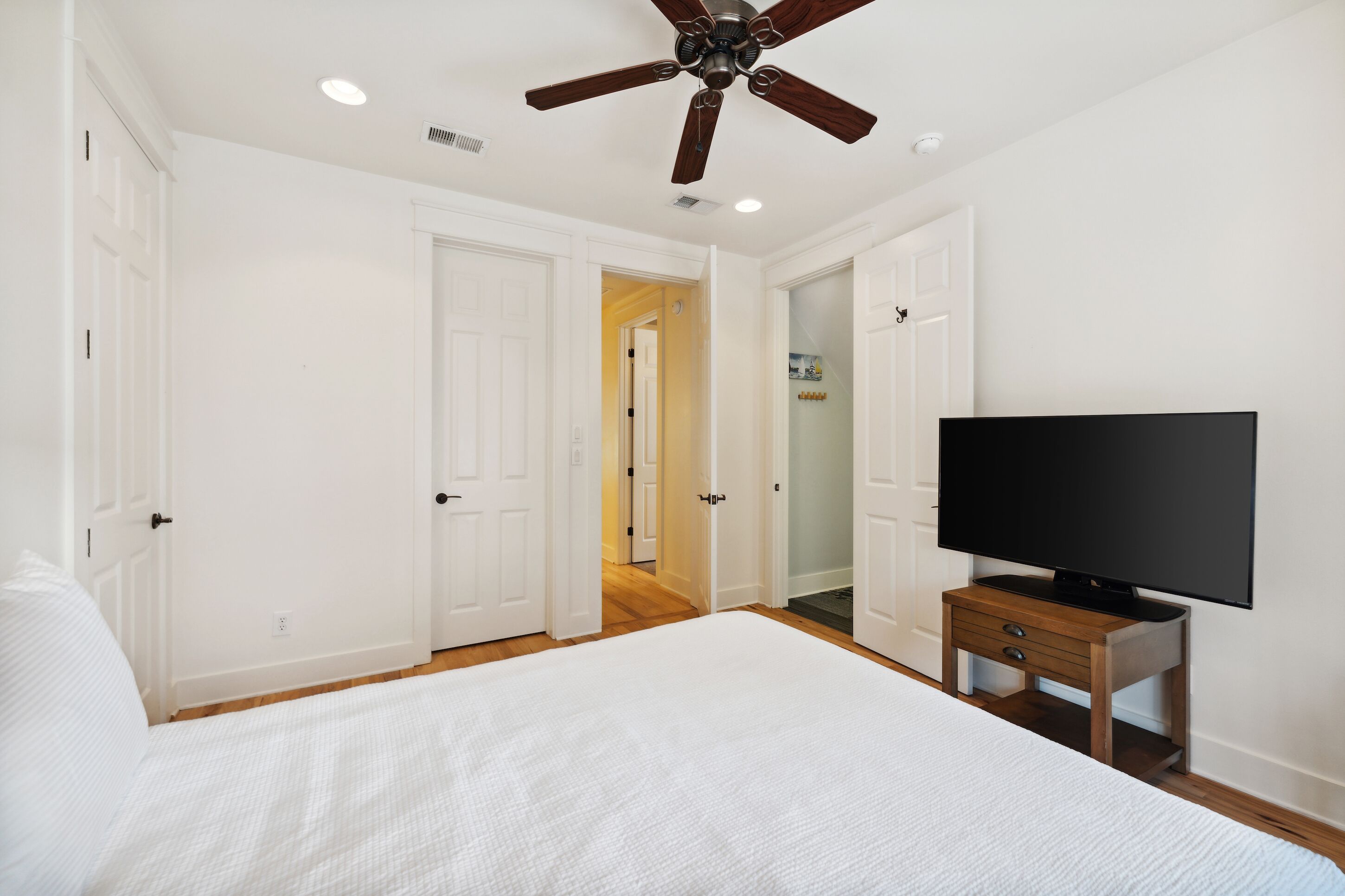 Un dormitorio con una cama grande, un ventilador de techo, un televisor sobre un soporte de madera y puertas blancas que dan a otras habitaciones.