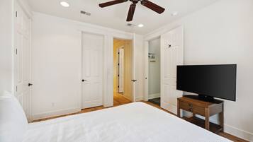 Un dormitorio con una cama grande, un ventilador de techo, un televisor sobre un soporte de madera y puertas blancas que dan a otras habitaciones.