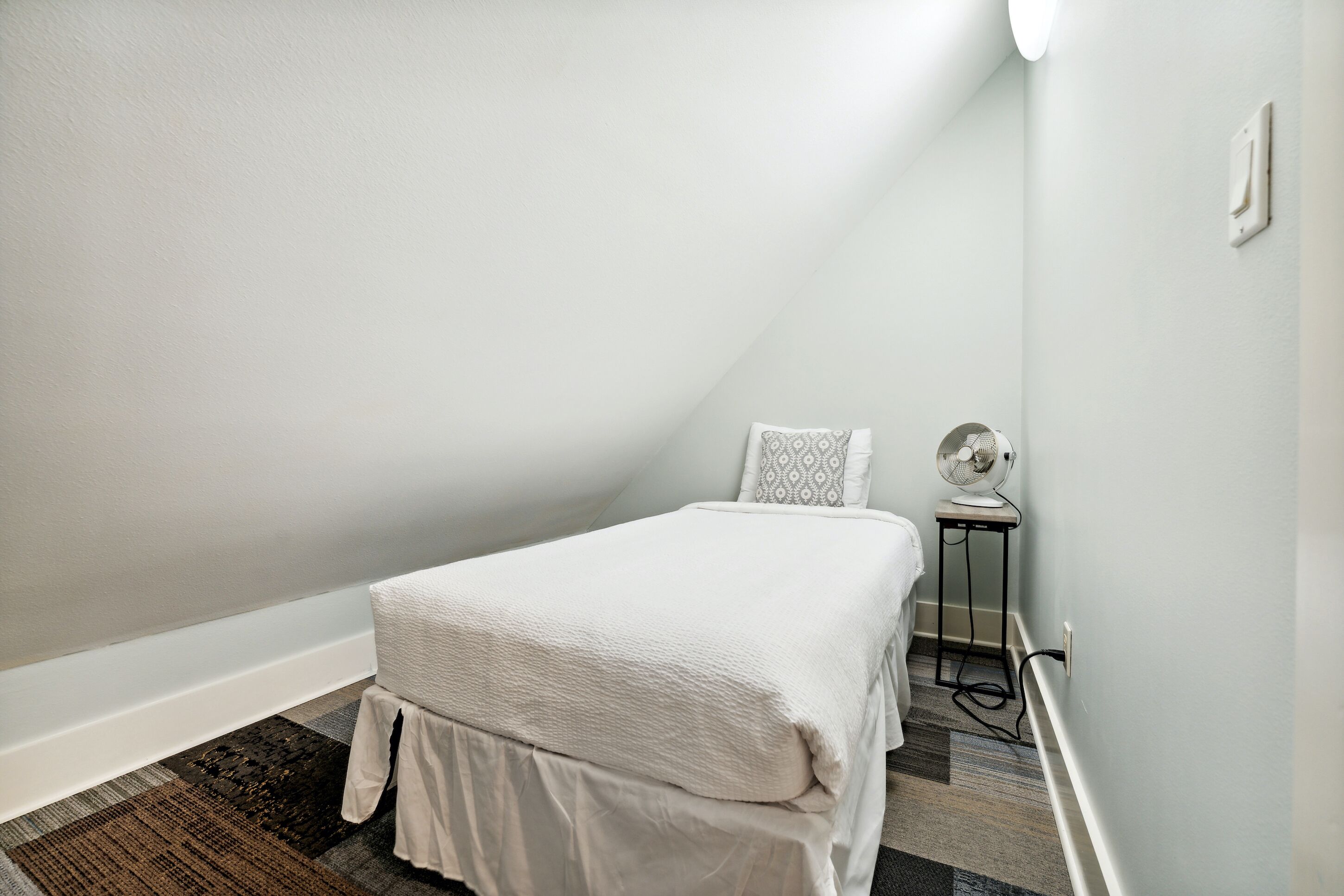 A small, neatly arranged bedroom with a bed, a bedside table, and a wall-mounted fan.