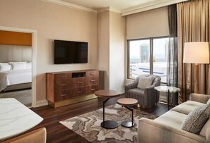 Une chambre d’hôtel comprenant un lit, une télévision, un coin salon avec un canapé et des fauteuils, une petite table et une grande fenêtre donnant sur des immeubles.