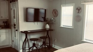 A bedroom with a bed, a desk with two stools, a ceiling fan, and a TV on the wall.