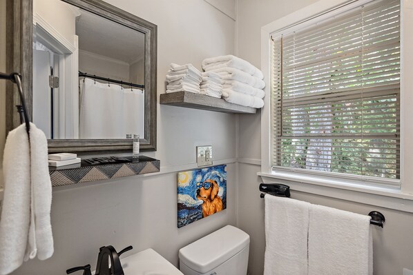 A bathroom with a toilet, sink, mirror, towel rack, and a window with blinds.