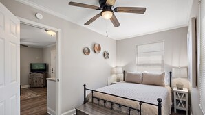 A bedroom with a bed, bedside tables, a ceiling fan, and a television in the adjacent room. - Cottage 4 - Nobody Inn (Ridgeland)