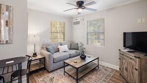 A living room with a grey sofa, a coffee table, a TV on a wooden cabinet, a ceiling fan, and a window with blinds. - Cottage 4 - Nobody Inn (Ridgeland)
