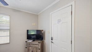 A room with a television on a wooden cabinet, a white door, and a window with blinds. - Cottage 4 - Nobody Inn (Ridgeland)