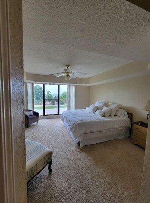 Relax in this serene bedroom with a large bed, nightstand, and chair, surrounded by natural light and a ceiling fan.