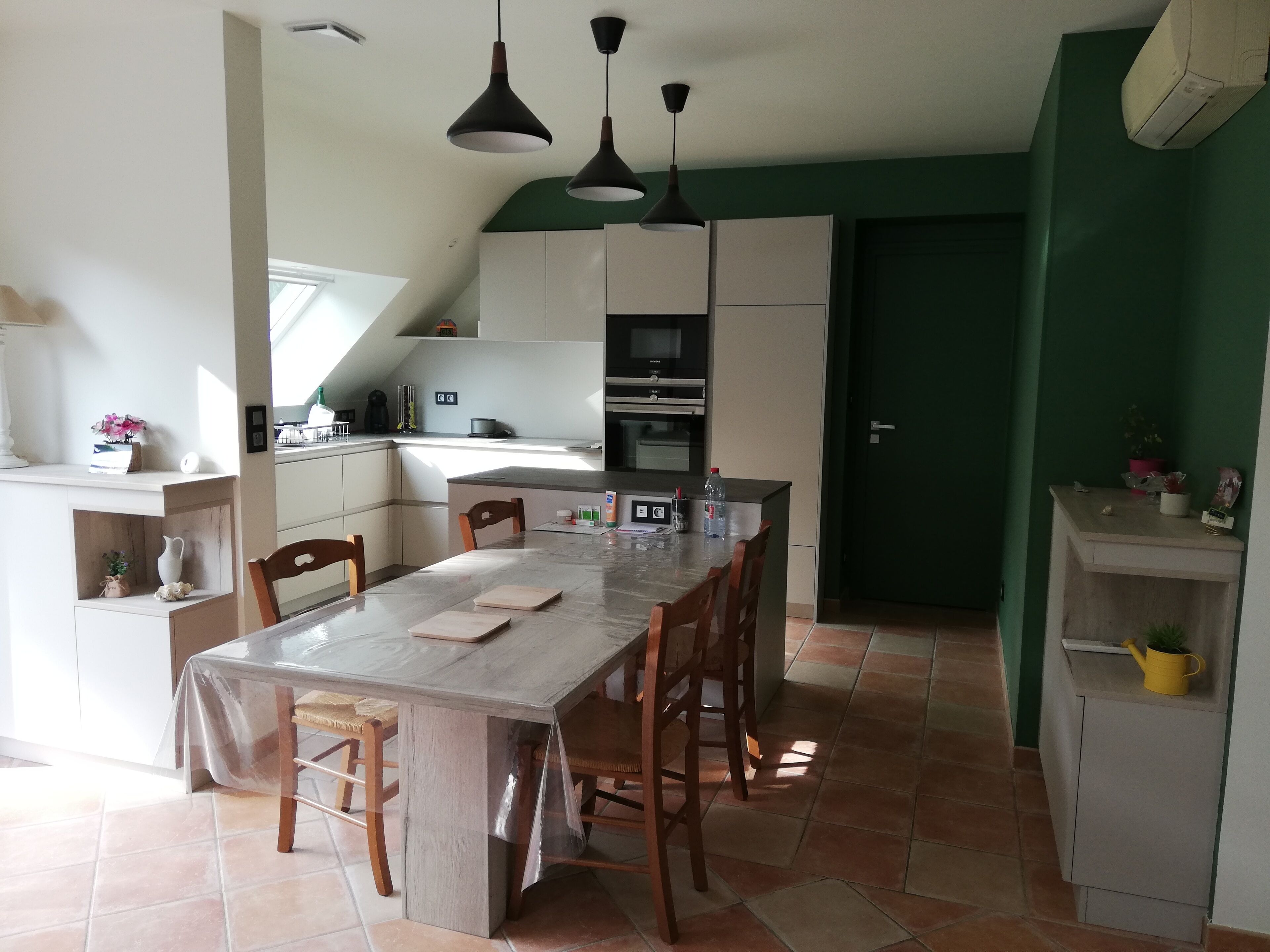 Een keuken met een eettafel, stoelen en hanglampen. Achtergrond is een groene muur en een deur.