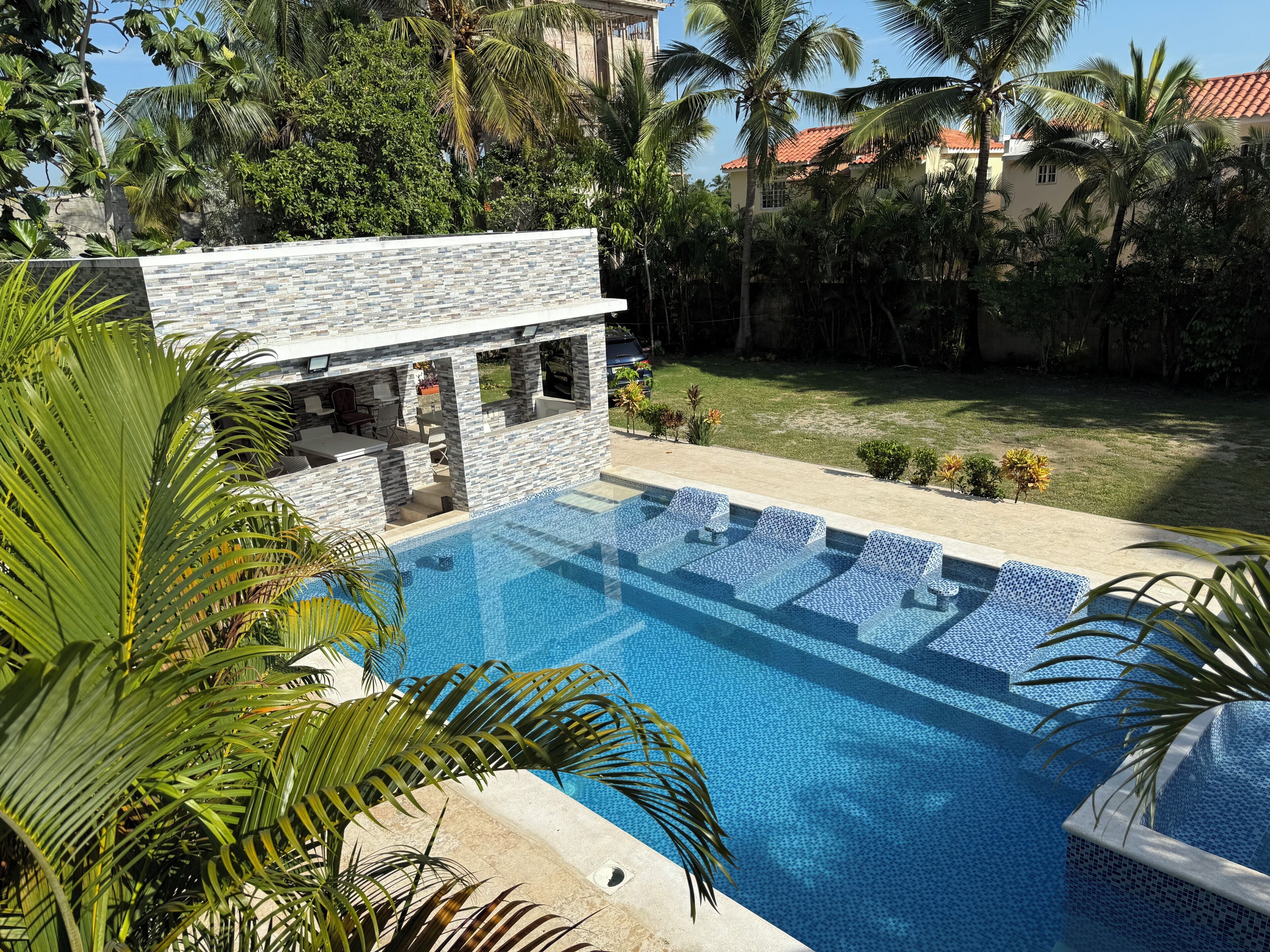 A swimming pool with a stone wall and lounge chairs, surrounded by palm trees and a grassy area.