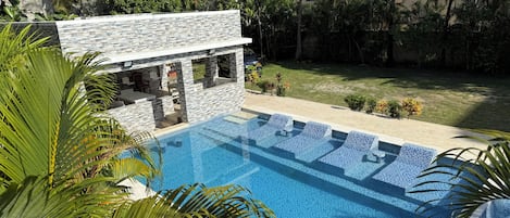 A swimming pool with a stone wall and lounge chairs, surrounded by palm trees and a grassy area.