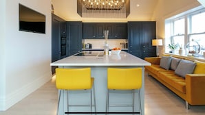 A modern kitchen with dark cabinets, a central island with two yellow bar stools, and a large chandelier. - Executive 3 bedroom Penthouse apartment, 2 bath and Mezzanine (Harrogate)