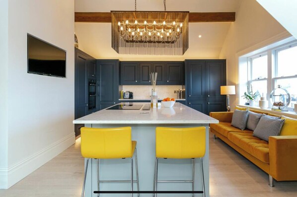 A modern kitchen with dark cabinets, a central island with two yellow bar stools, and a large chandelier.