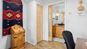 A room with a wooden cabinet, a red and blue patterned rug, a wooden door, a kitchen area with wooden cabinets, and a wicker basket on the wall. - Bosque "Abeyta" Casita (Bosque)
