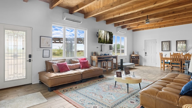 A spacious living room with a high ceiling, a large window, a sofa, a coffee table, and a dining area. - Bosque "Abeyta" Casita (Bosque)