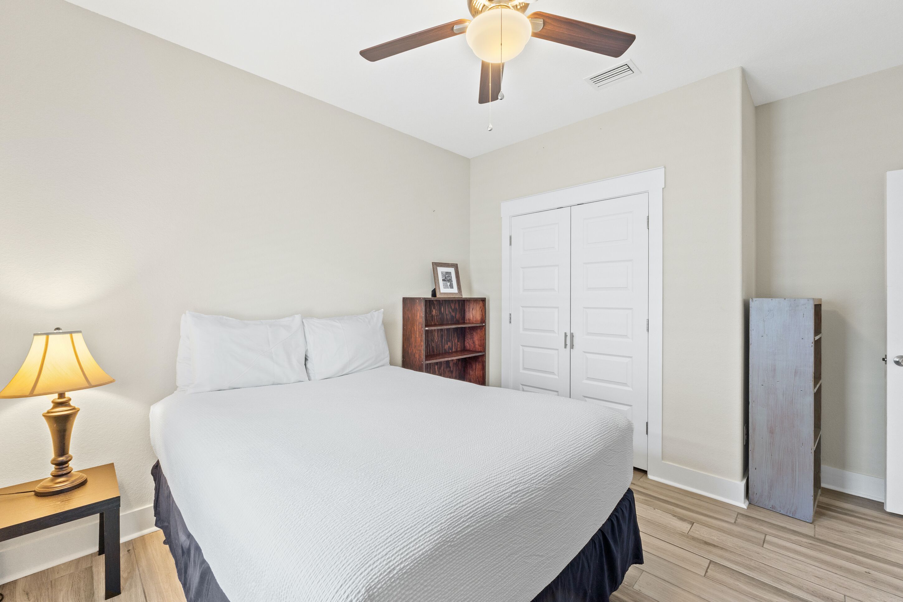 A bedroom with a bed, a nightstand with a lamp, a wooden bookshelf, a wardrobe, and a ceiling fan.