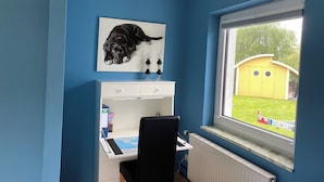 A room with a desk, a chair, a radiator, a window with a view of a garden, and a painting of a dog on the wall.