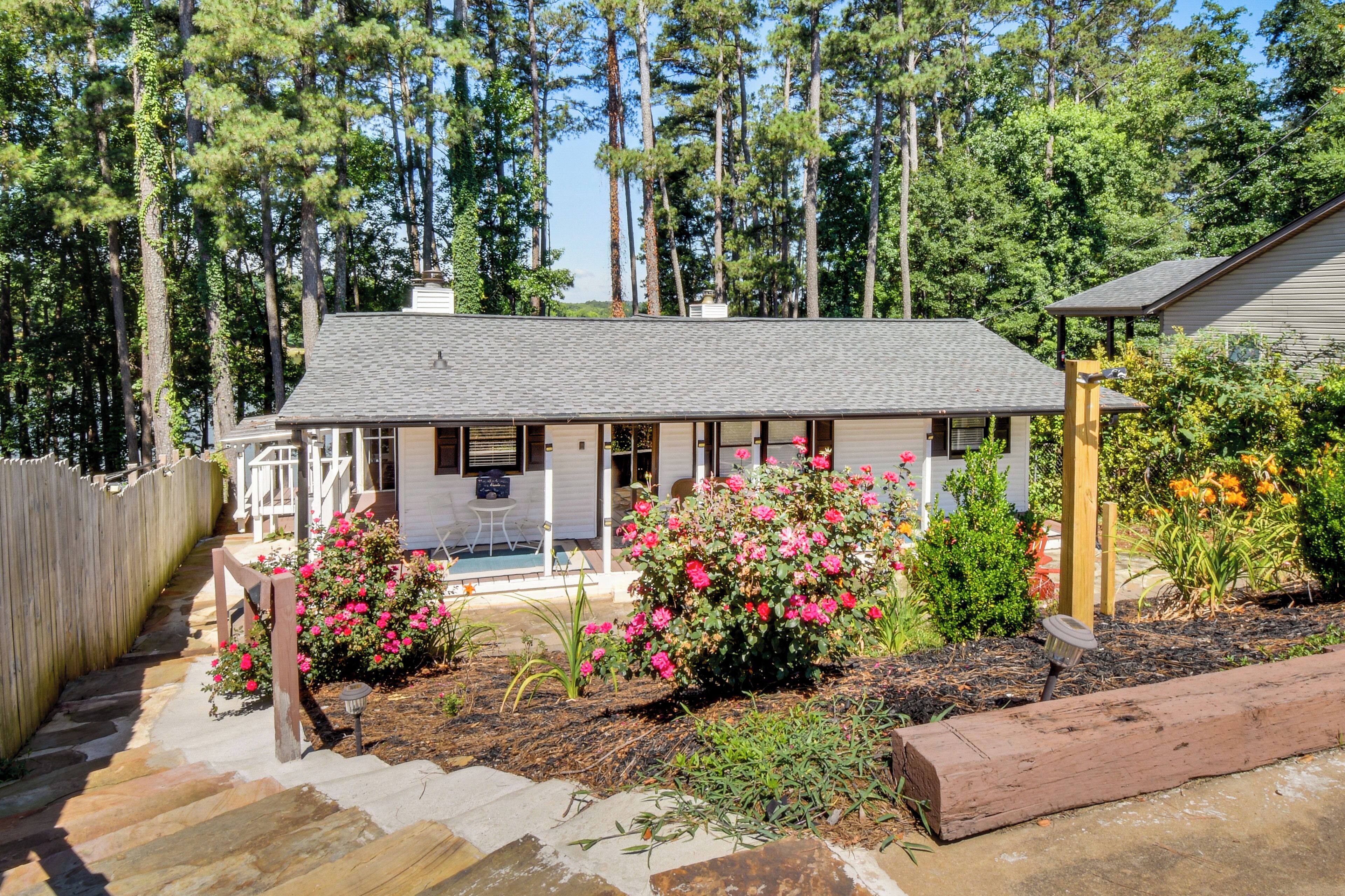 Uma casa com varanda, um pequeno jardim com flores cor-de-rosa e um deck de madeira.