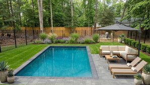 A rectangular swimming pool with a surrounding patio area, a wooden deck, and a seating arrangement. - Tranquil Modern Farmhouse (Waldorf)