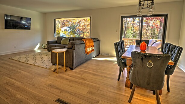 A modern living room with a grey sofa, a dining table with chairs, and large windows with autumn foliage views. - Tranquil Modern Farmhouse (Waldorf)