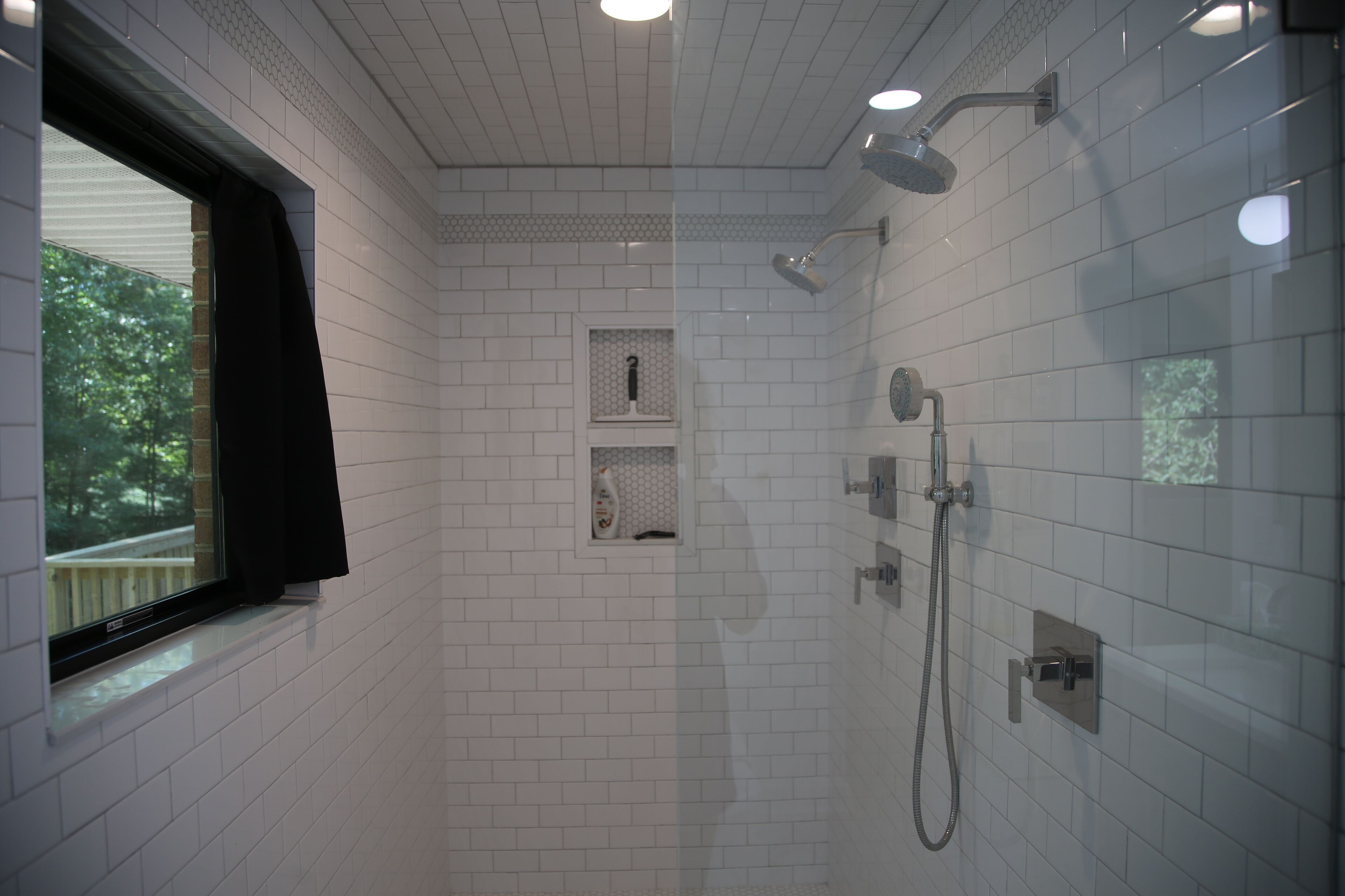 Un bagno con pareti piastrellate in bianco, una finestra con tenda nera, una doccia con soffione manuale e un piccolo ripiano con articoli da toilette.