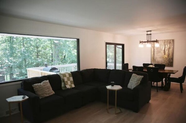A modern living room with a black sectional sofa, a small side table, a dining area with a wooden table and chairs, and a large window offering a view of trees and a building.
