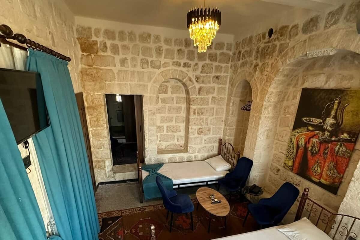 Ein Zimmer mit Steinwand, einem Bett, einem Schreibtisch, zwei Stühlen, einem kleinen Tisch und einer Chandelüster.