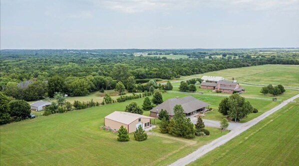 An aerial view of a rural property with multiple buildings, including a large barn and smaller structures, surrounded by expansive green fields and dense forests.
