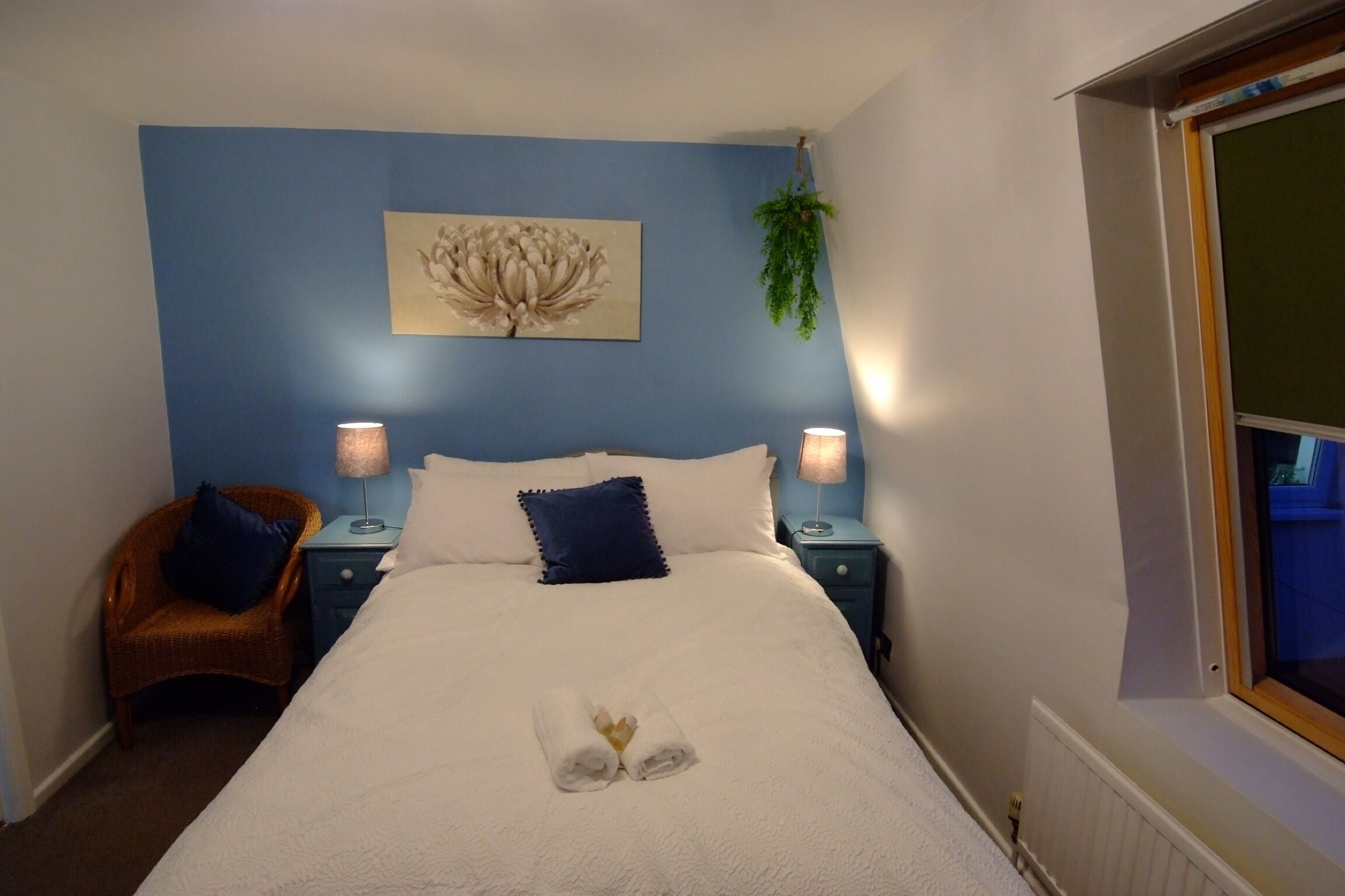 Un lit bien fait, avec une serviette et un objet décoratif posés dessus, un mur bleu orné d’un tableau floral, deux tables de chevet avec des lampes et une fenêtre offrant une vue agréable.