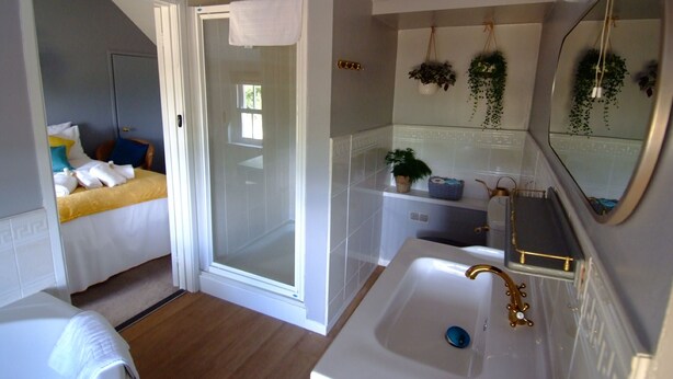 Habitación de hotel compacta con ducha, una cama con ropa de cama blanca y amarilla, un lavamanos blanco con detalles dorados y un espejo con una planta colgante.