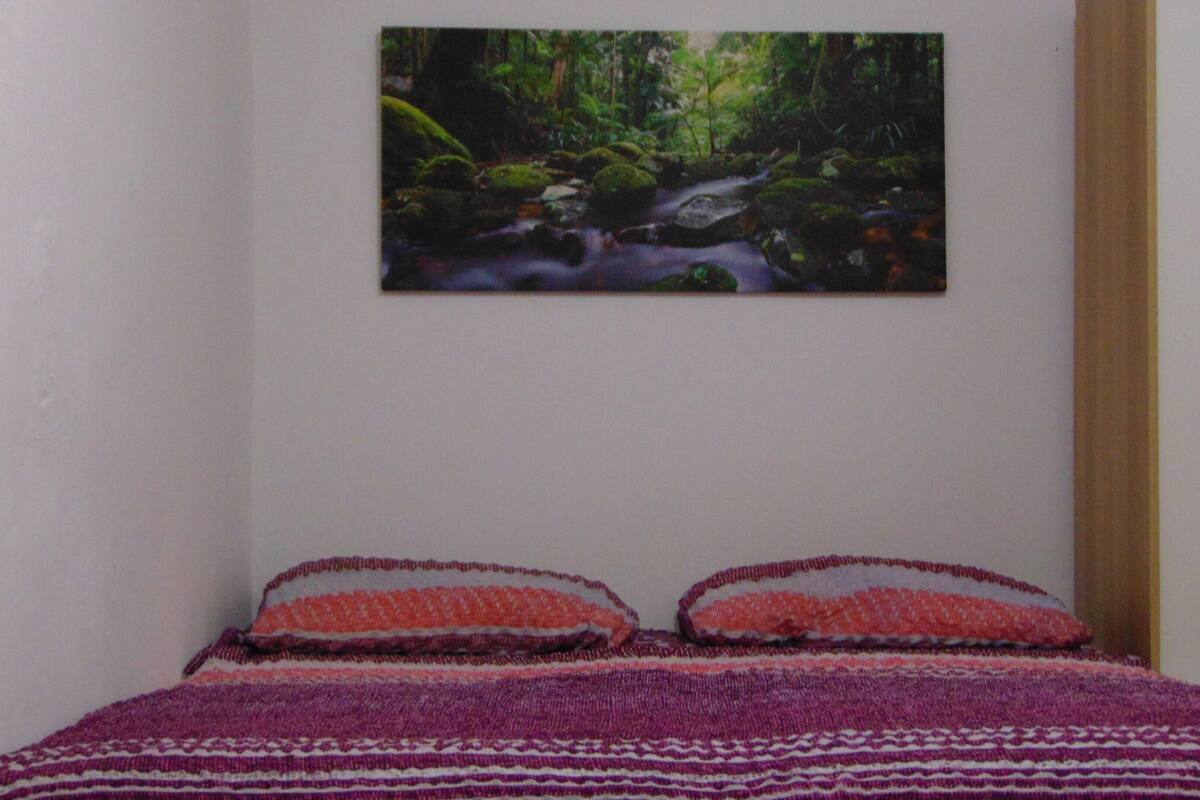 A bedroom with a bed, a painting of a forest stream on the wall, and a wooden door.