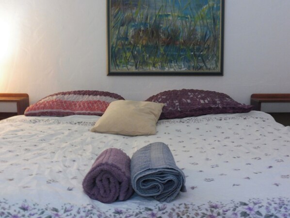 A neatly made bed with a floral-patterned bedspread, two rolled towels, and three decorative pillows.