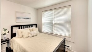 Une chambre à coucher comprenant un lit, une table de chevet, une fenêtre avec des stores, un luminaire au plafond et un tableau accroché au mur.