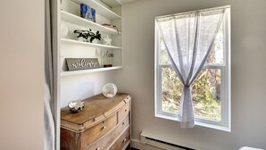 Une pièce avec un mur blanc, une fenêtre ornée de rideaux légers, une commode en bois agrémentée d’objets décoratifs et des étagères où sont disposés divers objets, dont une pancarte indiquant « Bienvenue ».