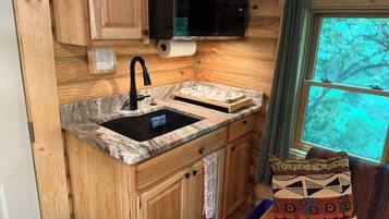 A compact kitchen with wooden cabinetry, a black sink, a microwave, and a window with a view of trees.