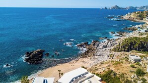 A coastal view with a rocky beach, clear blue sea, and buildings with balconies overlooking the ocean.