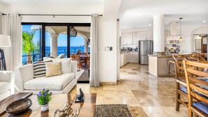 A modern living room with a view of the ocean, a kitchen with white cabinets, and a dining area with wooden furniture.