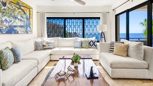 A modern living room with a large sectional sofa, a coffee table with decorative items, a ceiling fan, and a view of the ocean through large windows.