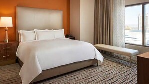 Habitación de hotel con una cama grande, una lámpara de noche, un banco y vistas a la ciudad a través de la ventana.