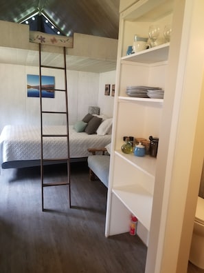 Un dormitorio con literas, una escalera, estante con utensilios de cocina y un cuadro en la pared.
