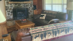 A cozy living room with a stone fireplace, a flat-screen TV mounted on the wall, a leather sofa with a decorative throw, and a wooden coffee table. - Cabin close to Helen and Water (Hiawassee)