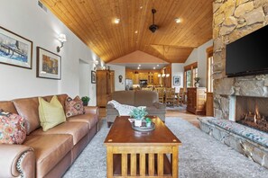 A cozy living room with a stone fireplace, wooden ceiling, and a dining area in the background.