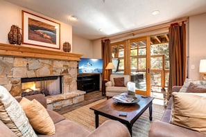 A cozy living room with a stone fireplace, comfortable seating, and a large window offering a view of the outdoors.